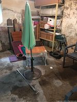 Green fabric outdoor umbrella closed and attached to a circular black metal base, next to a small metal side table with a wood top on a basement floor.