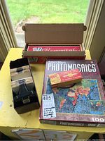 View of Rummikul box, Photomosaics puzzle box, Yogo Jr. game box stacked on yellow table with a green background.