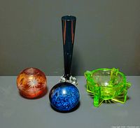 All four glass items arranged on a flat gray surface: cranberry glass vase, blue cut glass vase, blue glass paperweight, uranium glass ashtray