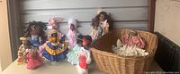 Wide view of 7 handmade dolls displayed beside a woven basket containing wrapped dolls or doll parts on a table.