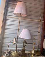 Five lamps arranged together against a metallic storage wall, showing different styles including floor and table lamps with shades and a silver desk lamp.