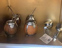 Four mate gourds with metal rims and medallions, three having metal straws, placed on a shelf with two new steel scoops.