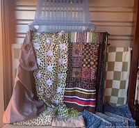 Rack with blue Rubbermaid basket on top, green painted metal rack, assorted curtains and linens draped over and hung on the rack including handmade quilt and table runner.