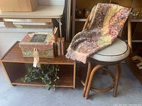 Vintage rattan swivel chair with white cushion with brown trim, decorative box, faux floral greenery and books on and around wooden shelf and chair.
