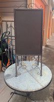 Two-panel metal chalkboard sign standing on ornamental wrought iron scrollwork metal stand placed on the round table.