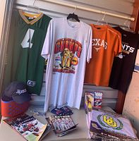 Four sports T-shirts and jersey hanging on rod with sports magazines and VHS tapes displayed below. Various team memorabilia including Houston Rockets NBA championship T-shirt, Green Bay Packers Brett Favre jersey, Texas Longhorns and New Orleans Saints T-shirts, Rice Owls cap, magazines and VHS tapes.