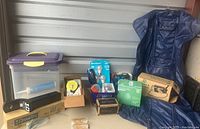 Overview of lot showing plastic storage bin, vintage GE iron in box, face masks, lightbulbs, Swingline staples, boxed 24-hour dial time switch, Draw-Tite ball mount kit box, and vintage metal ironing board with blue covering.