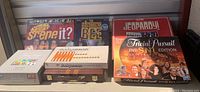 Wide view of five games including Jeopardy! metal case, SNL Trivial Pursuit DVD, Singing Bee, Seinfeld Scene It? DVD, and wooden Backgammon set.