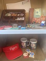 Shelf showing a brown compartment tray with pins and collectibles, faux money napkins, and a colorful Jumi cow figurine with tags. Also includes some sports memorabilia like a red Tigers visor and mugs with logos.