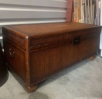 Full view of rattan and wood storage trunk with bun feet and metal handles on sides.