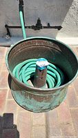 Brass garden hose container with an aged green patina holding a coiled green garden hose, placed against a white wall and connected to outdoor water spigots.