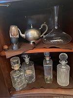 Photo of shelf showing various glass decanters, one metal tea kettle with white handle, and a glass wine cork stopper.