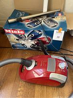 Photo showing red Miele canister vacuum cleaner with hose, alongside the original Miele box depicting various vacuum models.
