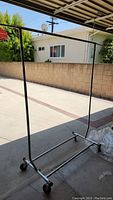Single folding portable metal clothing rack with horizontal hanging bar, vertical supports, and base with caster wheels, photographed outside.