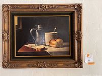 Full view of framed oil painting hanging on wall, depicting pewter pitcher, bread loaf, bowl and spoon still life