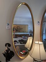 Front view of the large oval mirror showing the gold trim and reflective surface with furniture reflected in background.