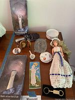 Overview of lot items on wooden table including Tiffany bowl, doll, decorative plaques, magnifiers, trinket dish, and porcelain spoon.