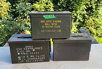 Three metal military-style ammunition boxes stacked outdoors on a wooden surface, two with visible stencil markings, one without markings.