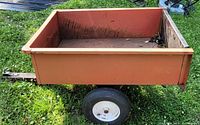 Front view of orange metal lawn trailer showing dumping bed and rubber tires.