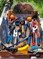 Overview of lot showing various hand tools and saw blades arranged in box outdoors under sunlight.