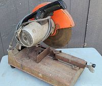 Full side view of the Makita steel chop saw, showing the heavy rusted metal base, clamp assembly, motor housing, and orange blade guard.
