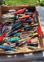 Wide view of assorted screwdrivers in cardboard box on outdoor table showing variety of handle styles and colors.