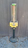 Full view of vending machine showing tall cylindrical coin rack, yellow vending box, coin slot, and black metal pedestal stand on gravel ground against dark wall.