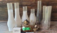 Overview of lot showing seven frosted glass chimneys, five clear glass chimneys, amber glass finger lamp with metal burner, and two additional burners.