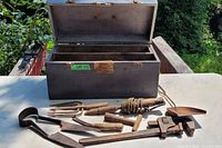 Open wooden toolbox with various tools laid out in front on a table outside. Tools include shears, pipe wrench, hammer, hand cultivator fork, and other wooden-handled metal tools. The toolbox shows visible signs of use and age.