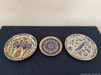 Three enamelled metal plates laid flat on dark surface, showing colorful rooster, blue and white symmetrical pattern, and blue and white bird designs.