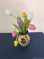 Front view of antique Gouda vase filled with 7 colorful German glass tulips on a dark surface against white background.