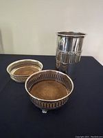 Photo of three silver-plated wine service pieces: two round coasters with pierced sides and felt pads, and one tall cylindrical bottle holder with pierced decorative band.