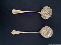 Photo of two silver berry serving spoons displaying front side, one with scalloped floral pierced bowl and plain handle, the other with round feather patterned pierced bowl and floral engraved handle, on black background.