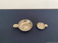 Two silver tastevins (wine tasting cups) of differing sizes with small flat handles, placed side by side on a dark surface.