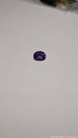 Oval cut deep purple Bolivian amethyst gemstone lying on white background, showing top view of facets and color saturation.