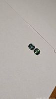 Two small oval cut sapphires in multicolored ice blue and green hues on a neutral background.