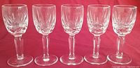 Five clear Waterford Irish Crystal cordial glasses in Lismore pattern on a red background.