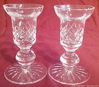 Pair of clear Waterford Irish Crystal single light candlesticks with diamond cut pattern on body and sunburst cut on base, displayed on red fabric background.