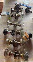 Top-down view of all six door knob sets including clear and black glass knobs plus brass hardware and metal hinges grouped on a table surface