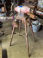 Full view of heavy industrial grinder mounted on rusty tripod metal base with pink metal housing and power cord.