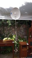 Front view of the tall floor lamp with white parabolic metal shade and exposed light bulb, standing outdoors against a stone wall with greenery.