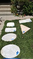 Photo showing six white marble slabs of various shapes (oval, rectangular, triangular) laid out on grass near stone steps, with blue tape labels on each slab.