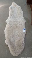 Single photo of an off-white irregular shaped sheepskin rug laid flat on a gray concrete floor, showing its texture and shape with a small tag attached.