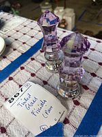 Pair of lilac tinted glass finials placed on a patterned cloth showing the front and side view of the pieces alongside a tag noting 'Tinted Glass Finials Pair'