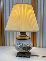 Full view of bedside table lamp with pleated fabric shade illuminated, showing lamp base and porcelain center with floral design.
