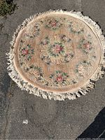 Overhead image showing full round rug with floral pattern and fringed edges, placed outdoors on asphalt surface