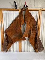 Full front view of two brown leather western cowboy chaps hanging with fringe details and leather lower legs.