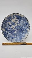 Front view of round scalloped porcelain plate with blue hand-painted peacock and floral design and measuring ruler for scale