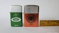 Front view of two collectible lighters, one green with 'Minnesota Rubber' logo and one copper with 'Jet Commander Around the World' logo, placed beside a measuring ruler indicating size.