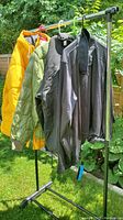 Four men's jackets hanging on a rack outdoors in sunlight: yellow puffer, green quilted, black zip jacket, and black pullover